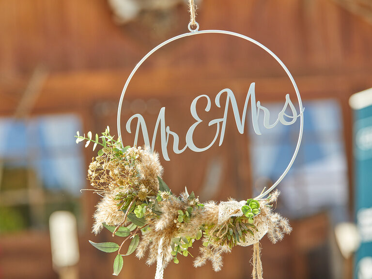 Detailaufnahme eines 'Mr & Mrs' Hochzeitsdekors mit getrockneten Blumen im Hintergrund einer Hütte.