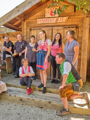 Gruppe fröhlicher Menschen in traditioneller Tracht vor einer Holzstruktur die im Freien zusammenarbeitet und sich unterhält.