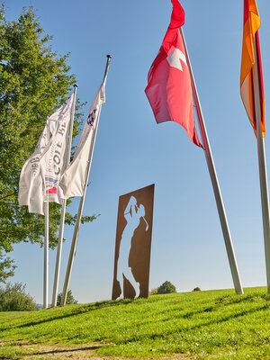 Golfer beim Abschlag auf einer Anhöhe, umgeben von wehenden Fahnen und einer stilisierten Golfer-Skulptur.