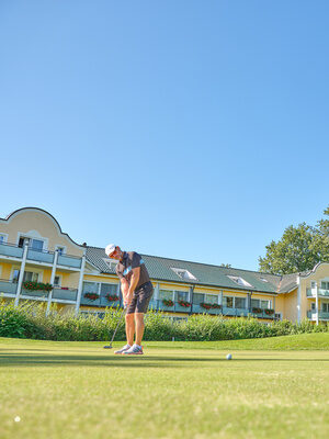 Golfer beim konzentrierten Putt auf dem Grün vor einem stilvollen Hotel mit Balkonen.