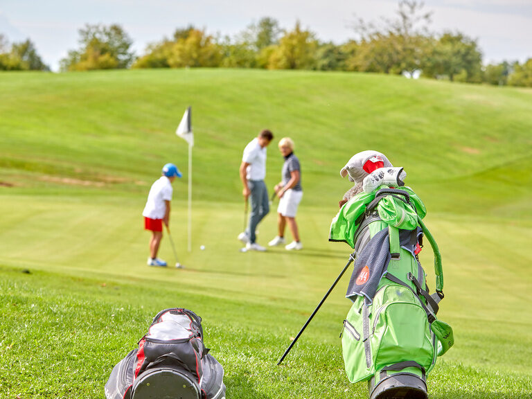 Golfbags im Vordergrund, während ein Kind und Erwachsene üben – perfektes Ambiente für einen Golfkurs bei Bad Griesbach.