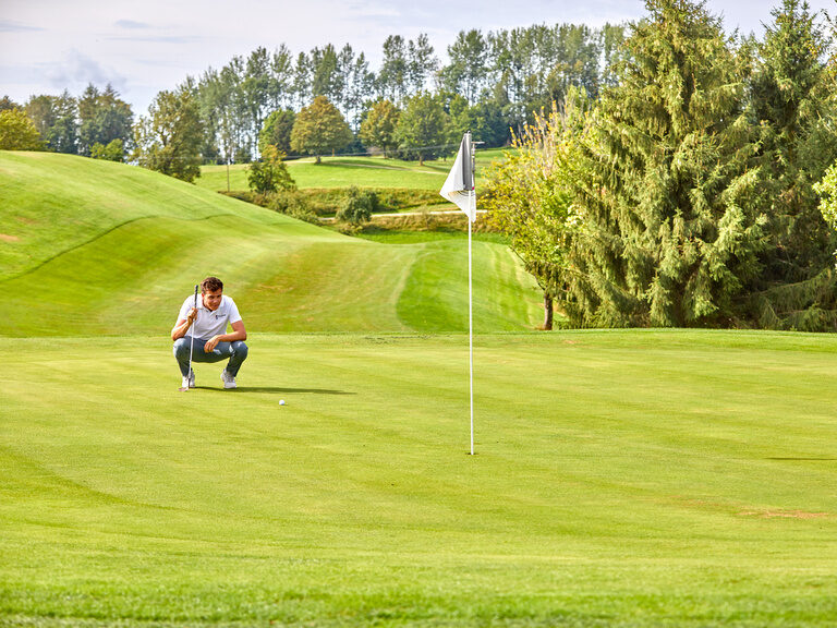 Ein Golfer, der sich auf dem Grün konzentriert, mit Fahne im Vordergrund und einer idyllischen Kulisse aus Bäumen und Wiesen.