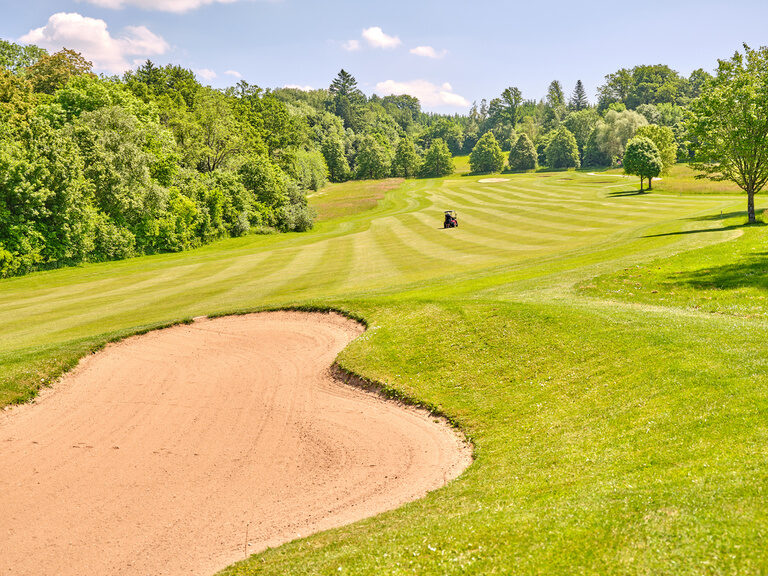 Ein weitläufiges Fairway mit einem Sandbunker und einem Golfcart, eingebettet in eine grüne, bewaldete Landschaft.