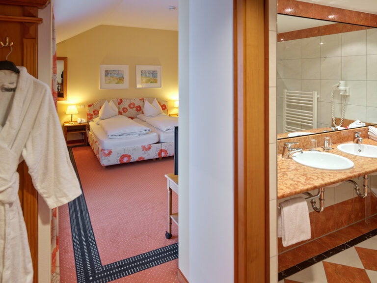 Hotelzimmer mit zwei Betten in floraler Optik und Blick ins Badezimmer mit großem Spiegel und Doppelwaschbecken.