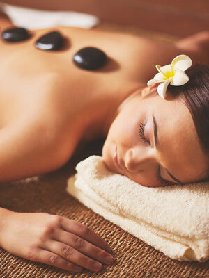 Frau bei einer Hot-Stone-Massage auf einem Handtuch, entspannt mit Blüte im Haar in einem warmen Raum.
