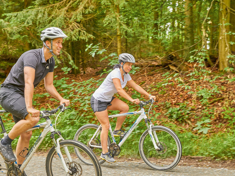 Zwei Radfahrer genießen einen Ausflug auf einem schattigen Waldweg während ihres Radurlaubs in Bayern.